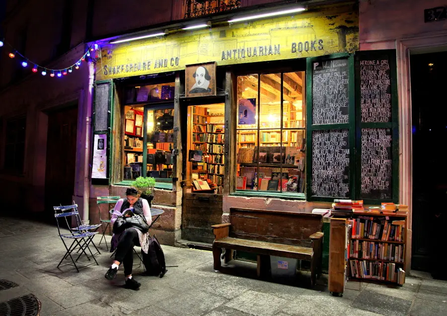 A reader sits outside Shakespeare and Company bookshop in Paris at night, its windows glowing with shelves of books